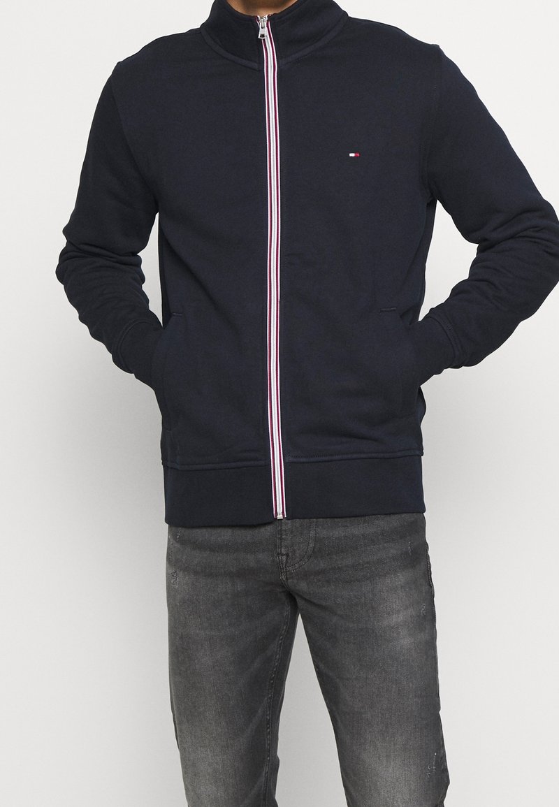 Granatowa bluza rozpinana na zamek z wysokim kołnierzem oraz kontrastowym zamkiem w czerwono-białe paski. Posiada dwie boczne kieszenie i małe logo na piersi.