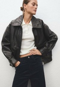 Črna usnjena bomberska jakna z širokim ovratnikom, rebrastimi manšetami in zadrgo spredaj, v kombinaciji z belim rebrastim zgornjim delom in temnimi hlačami.