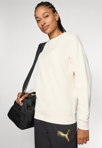 Cremfarbener Sweatshirt mit langen Ärmeln und einem kleinen Logopatch. Kombiniert mit dunklen Hosen mit goldenem Logo und einer schwarzen Tasche mit einem Tragegurt.