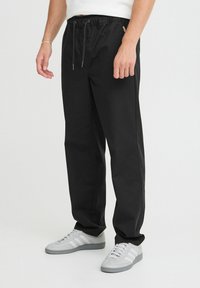 Sorte bukser med snore i taljen, tapered form og glat overflade, parret med grå sneakers med hvide striber.