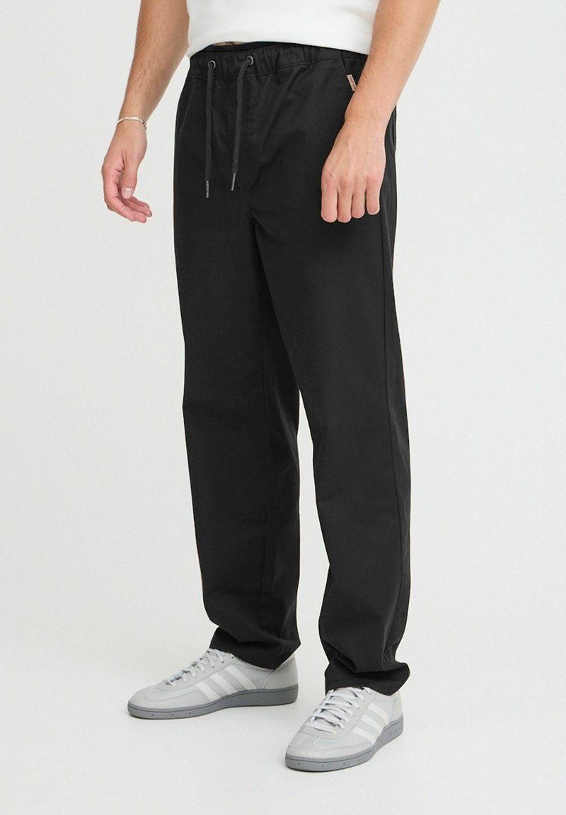 Sorte bukser med snore i taljen, tapered form og glat overflade, parret med grå sneakers med hvide striber.