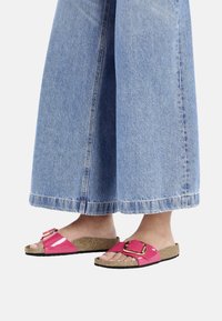 Infradito rosa lucide con una grande fibbia, soletta in sughero e suola nera, abbinate a pantaloni di jeans blu a gamba larga.