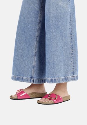 Roze glanzende slidesandalen met een grote gesp, kurken voetbed en zwarte loopzool, gecombineerd met wijde blauwe spijkerbroek.