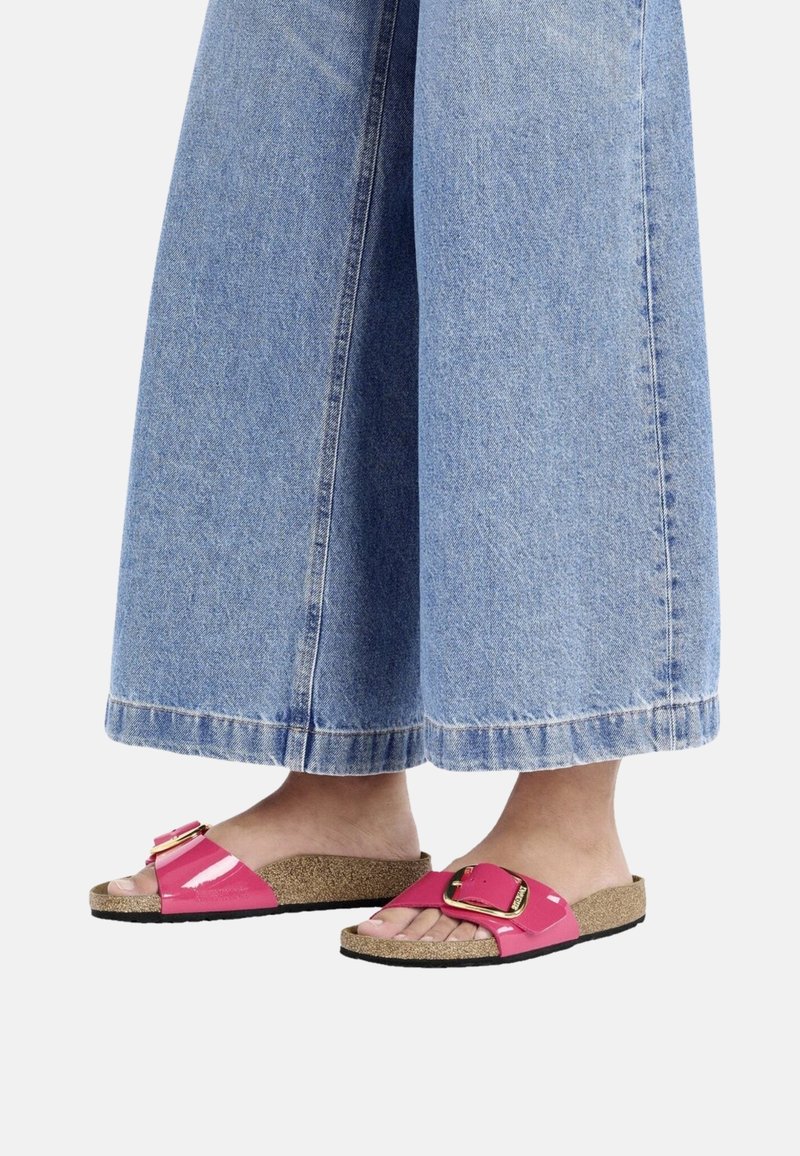 Infradito rosa lucide con una grande fibbia, soletta in sughero e suola nera, abbinate a pantaloni di jeans blu a gamba larga.