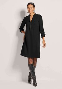 Schwarzes Kleid mit V-Ausschnitt und halben Ärmeln, ausgestattet mit einem Reißverschluss vorne und einer Brusttasche. Kombiniert mit knielangen schwarzen Stiefeln.