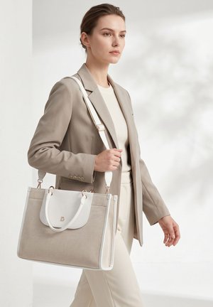 Jeune femme en blazer et pantalon beige porte un grand sac cabas beige et blanc avec une bandoulière, marchant à l'intérieur.