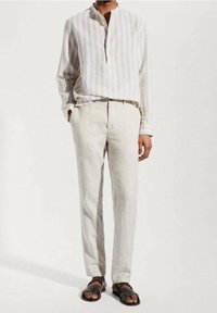 Persona vestida con una camisa de rayas beige de mangas largas, pantalones beige claros y sandalias oscuras de punta abierta, de pie contra un fondo claro y liso.