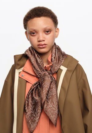 Joven con cabello corto y rizado que lleva una bufanda marrón con flores, camisa color coral y chaqueta marrón contra un fondo claro y liso.