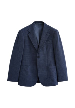 Junger Mann mit kurzen dunklen Haaren, der einen blau karierten Blazer über einem kastanienbraunen Hemd mit Knöpfen trägt und mit neutralem Gesichtsausdruck geradeaus schaut.