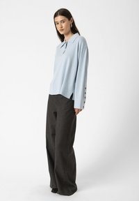 Woman wearing light blue long-sleeve knit top with buttons on cuffs and dark wide-leg trousers standing against a plain background.