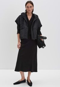 Gilet rembourré noir avec capuche, manches courtes et poches zippées, superposé sur un haut noir et une jupe longue. Les chaussures présentent des pointes et des rivets.