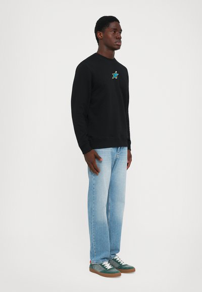 Man standing wearing a black sweatshirt with a blue star emblem, light blue jeans, and green suede sneakers with white laces.
