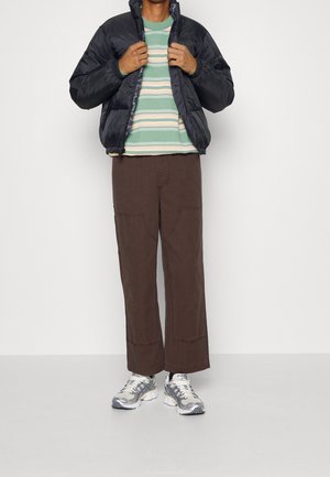 Černá prošívaná bunda, zelené a pruhované tričko s dlouhým rukávem, hnědé kalhoty s širokými nohavicemi a šedé tenisky s reflexními prvky.