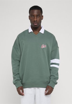 Homme portant un pull vert avec un col blanc et des rayures sur les manches, debout devant un fond uni gris clair.