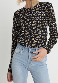 Haut à manches longues à imprimé floral sur fond noir, orné de fleurs jaunes et blanches. Associé à un jean taille haute bleu clair.
