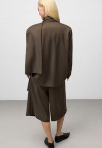 Frau mit langen blonden Haaren, die einen übergroßen braunen Blazer und passende weite knielange Shorts trägt, schwarze spitze Ballerinas, stehend auf grauem Boden.