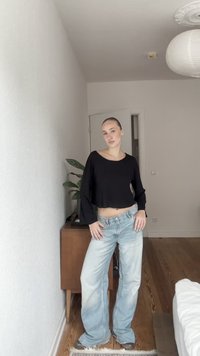 Svart kortermet genser med lange ermer, lys blå vide jeans med falmet utseende, brukt med grå sko, satt mot en enkel interiørbakgrunn.