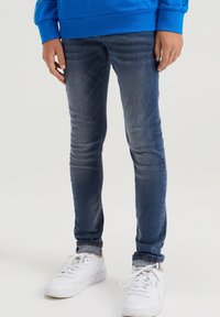 Jeans de denim azul con un ajuste slim, con un lavado desgastado y sutiles arrugas. Combinados con zapatillas blancas.