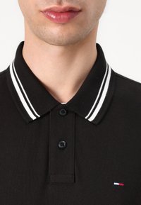 Čierna polo košeľa s golierom, s bielymi pruhmi na akcentoch a tromi čiernymi gombíkmi. Malé logo v červenej, bielej a modrej farbe na leme.