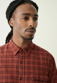 Punane ruuduline särk nööbid ees, krae ja kahe ees taskuga. Materjal on pehme tekstuuriga, mustade joontega ääristatud.