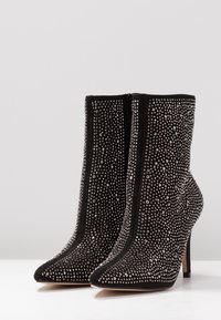 Botines de ante negros con puntas afiladas, tacones de aguja y cubiertos de pequeños tachuelas plateadas, posicionados uno al lado del otro sobre una superficie blanca.