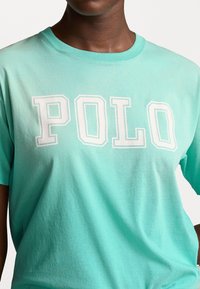 Tyrkysové tričko z měkké bavlny s krátkými rukávy a kulatým výstřihem, s bílými blokovými písmeny "POLO" přes hruď.