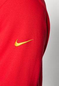 Raudonaablaudė su lygaus audinio, su auksiniu Nike logotipu kairiajame rankovėje. Paprastas dizainas su apvaliu iškirpimu.