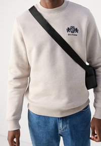 Beige sweatshirt with crew neck and ribbed cuffs, featuring a dark navy logo on the chest. Paired with blue denim jeans and a black crossbody bag.