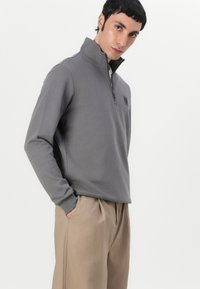Felpa grigia con zip e colletto alto, decorata con un logo ricamato, abbinata a pantaloni beige. Texture liscia e vestibilità casual.