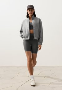 Femme marchant portant une veste grise zippée, un soutien-gorge de sport sombre, un short de vélo foncé, des chaussettes blanches, des baskets et une casquette de baseball sombre sur un sol clair.