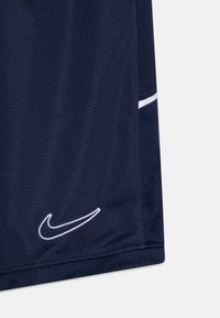 Tumsnā zila sporta šorti ar balto Nike swoosh logotipu izšūti pie apakšmalas un baltu horizontālu svītru uz acs sānu paneļiem.