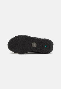 Suela de zapatilla negra con goma texturizada, con tracción en forma de triángulo y un logotipo central. Incluye un pequeño acento turquesa cerca del talón.