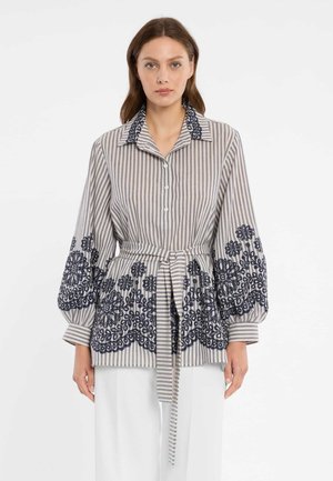 Vrouw draagt een beige gestreepte blouse met marineblauwe bloemenborduurwerk, pofmouwen, knopen aan de voorkant en een bijpassende stoffen ceintuur, gecombineerd met witte broek.