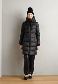Cappotto imbottito nero con chiusura a zip, cappuccio e lacci, caratterizzato da una silhouette lunga e aderente e una texture trapuntata lucida, abbinato a pantaloni neri.