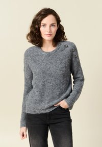 Maglione grigio lavorato a maglia con una trama morbida, scollo rotondo e bottoni decorativi sulla spalla sinistra. Abbinato a pantaloni in denim neri.
