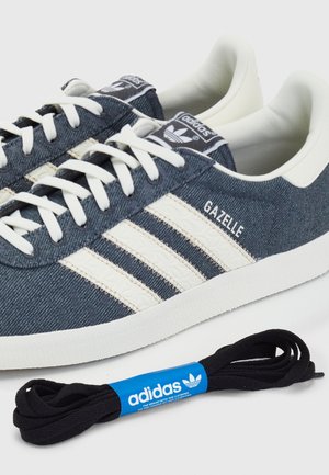 Niebieskie sneakersy Adidas Gazelle z białymi paskami i białymi sznurowadłami, pokazane z dodatkową parą czarnych sznurowadeł Adidas w opakowaniu.