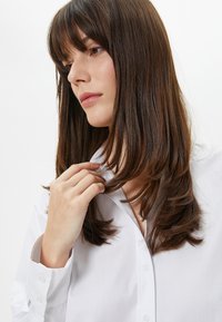 Camicia bianca con colletto e bottoni, texture liscia, tessuto morbido. Il soggetto tocca delicatamente i capelli. Lo sfondo è semplice e non invadente.