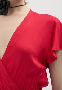 Primo piano di una persona che indossa un vestito rosso a V con maniche svolazzanti e una collana choker nera con un grande fiore di stoffa.