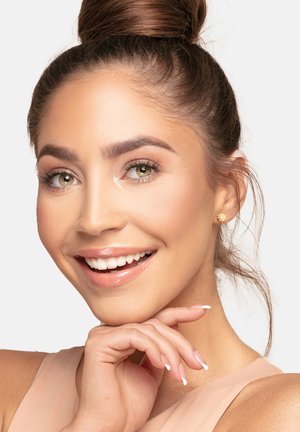 Eine Frau mit einem Dutt lächelt, trägt kleine goldene, blumenförmige Ohrringe, ein hellbeiges Oberteil und hat ein natürliches Makeup.