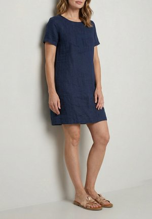 Kvinde iført en mørkeblå kortærmet hørkjole kombineret med roseguld flade slide sandaler, stående mod en lys væg.