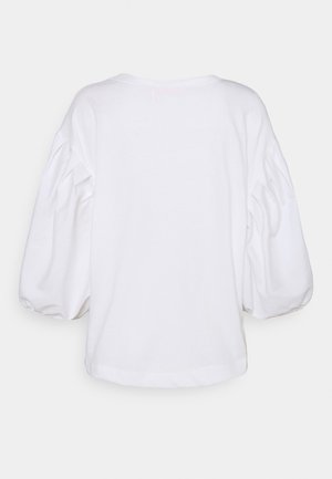 Nainen seisoo tumman seinän vieressä, päällään valkoinen liehukehihainen paita, laivastonsiniset napilliset shortsit, valkoiset lenkkarit ja hän pitää ruskeaa nahkaista säkkilaukkua.