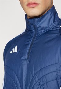Marineblaue Zip-Jacke mit hohem Kragen, die eine gesteppten Textur und weiße Logo-Details auf der Brust und Schulter aufweist.