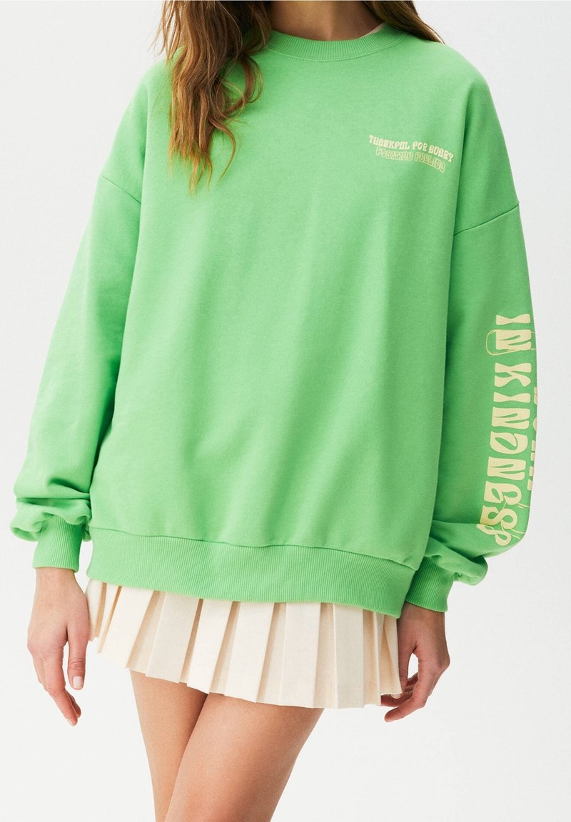 Femme portant un sweat-shirt oversize vert vif avec un texte jaune sur la manche et la poitrine, associé à une jupe plissée courte beige.