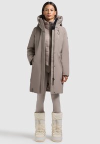 Beige isoleret parka med hætte, to forlommer og lynlåslukning, lagdelt over en matchende beige puffer vest og tætsiddende leggings. Hvide snesko.