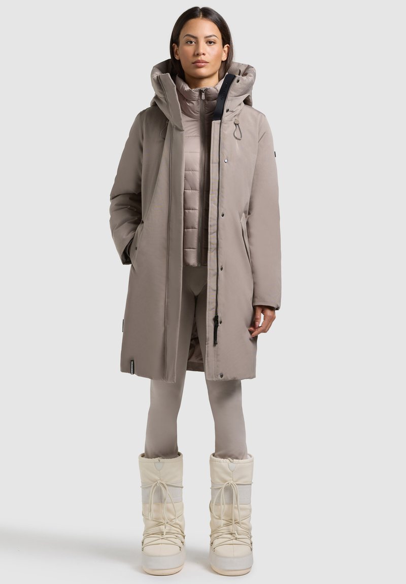 Beige isoleret parka med hætte, to forlommer og lynlåslukning, lagdelt over en matchende beige puffer vest og tætsiddende leggings. Hvide snesko.