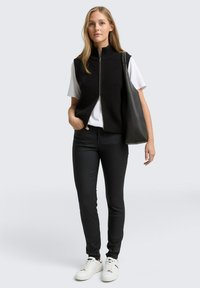 Svart ribbad stickad väst med hög krage, vit T-shirt, svarta åtsmitande byxor och vita sneakers, hållande en svart axelväska.