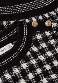 Pull en tricot à motifs noir et blanc avec deux boutons dorés en forme de tête de lion près du col et une étiquette de taille MNG visible.