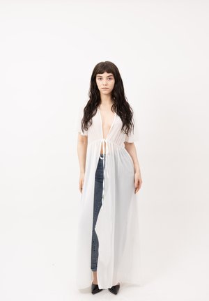 Vrouw met lang donker haar die een doorschijnende witte open voorkant badjas draagt, gestrikt in de taille, over een blauwe spijkerbroek en zwarte puntige schoenen.