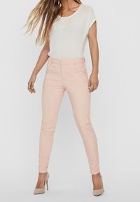 Světlé růžové úzké kalhoty s hladkým povrchem, předními kapsami a středně vysokým pasem, kombinované s volným bílým topem a béžovými podpatky.