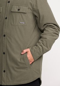 Veste chemise en velours côtelé vert olive avec des boutons de fermeture, deux poches poitrine et une coupe décontractée. Dotée d'un tissu texturé et de boutons noirs.